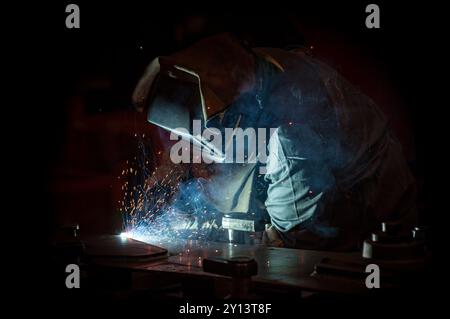 Welder on a black background. The light from the welding torch casts a bluish light that only illuminates the welder, in full PPE and his immediate wo Stock Photo
