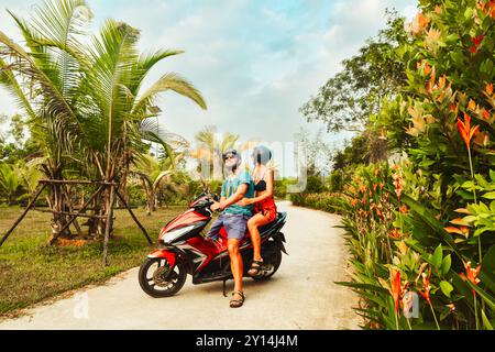 Funny authentic young happy caucasian couple pose on scooter together with palm trees, having fun driving red scooter on tropical road trip in southea Stock Photo