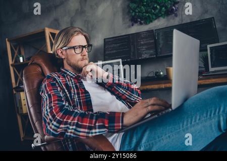 Photo of young blond bearded man thoughtful focused fixing code sitting leather boss armchair tapping laptop indoors loft modern office Stock Photo