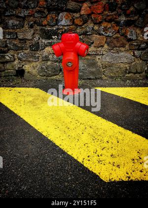 Bright red fire hydrant standing out against yellow lines on a textured stone wall in an urban setting during daylight Stock Photo