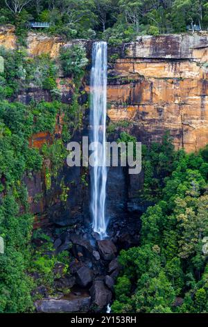 A tall waterfall flows smoothly over rugged cliffs, creating a beautiful display of water splashing onto large rocks below. The surrounding area is fi Stock Photo