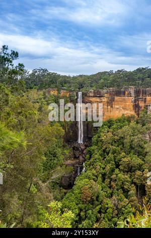 A tall waterfall flows smoothly over rugged cliffs, creating a beautiful display of water splashing onto large rocks below. The surrounding area is fi Stock Photo
