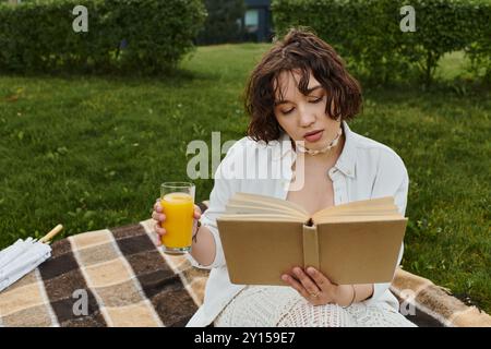 A young woman in a white shirt relaxes on a picnic blanket, savoring a drink while reading a book. Stock Photo