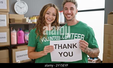 A smiling man and woman volunteers holding a 'thank you' sign in a donation center warehouse. Stock Photo