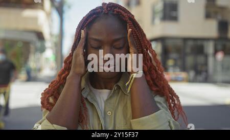 A serene young black woman with red braids standing on a city street, eyes closed, hands on temples, embodying tranquility amidst urban chaos. Stock Photo