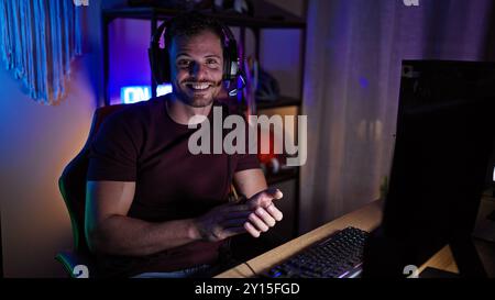 A smiling young hispanic man with a beard enjoying gaming in a dark room at night, creating a cozy and engaging atmosphere. Stock Photo