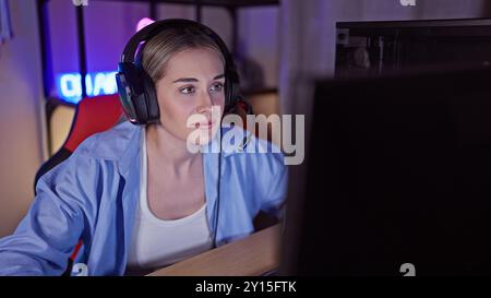A focused young woman wearing headphones engages in a gaming session indoors during nighttime. Stock Photo