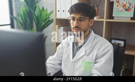 Arab man with beard in glasses, wearing a lab coat, works attentively at a computer in a hospital office. Stock Photo