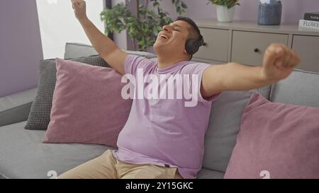A joyful hispanic man relaxes with headphones on a cozy sofa in a modern apartment. Stock Photo