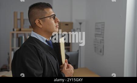 Hispanic man in a formal robe standing in an office, holding a folder with a focused expression, suggesting a legal professional such as a lawyer or j Stock Photo