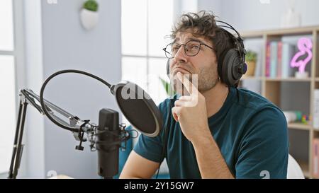 Pensive hispanic man with beard and headphones in radio studio contemplating while recording. Stock Photo