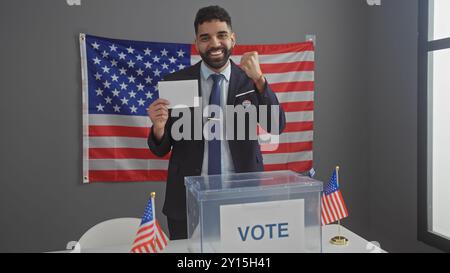 A smiling young hispanic man with a beard proudly showing an envelope while voting in an american electoral room with flags. Stock Photo