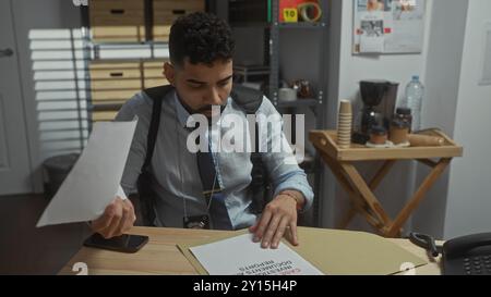 A focused hispanic man with a beard examines documents at a desk in a detective's office. Stock Photo