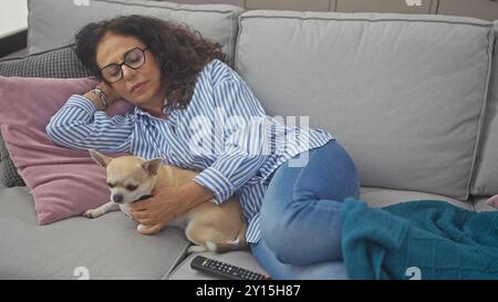 A middle-aged hispanic woman naps on a couch beside her chihuahua, in a cozy home setting. Stock Photo