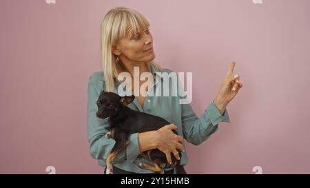 Mature blonde woman holding chihuahua dog while pointing finger on isolated pink background Stock Photo