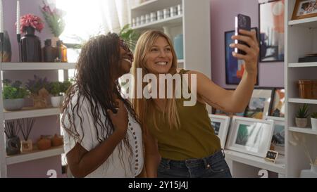Two women taking a selfie together in a home decor store, surrounded by decorative interior items, plants, and framed pictures on shelves. Stock Photo