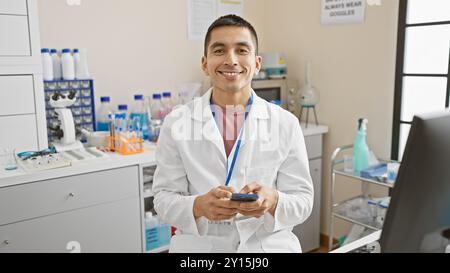 Hispanic man in lab coat smiling with smartphone in a modern laboratory Stock Photo