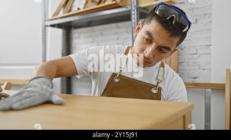 A focused hispanic man wearing safety glasses examines a wooden piece in a carpentry workshop. Stock Photo