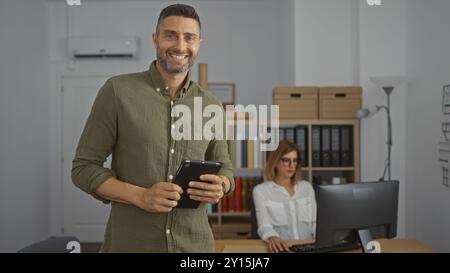 Man smiling with tablet standing in modern office while woman works on computer in background, showcasing professional business environment indoors Stock Photo