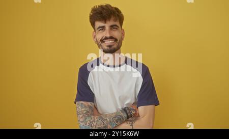 Handsome hispanic man with tattoos smiles confidently while crossing arms against a yellow background Stock Photo