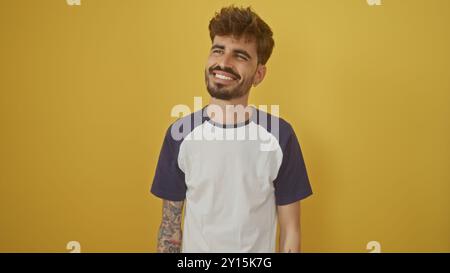 A handsome young hispanic man with a beard and tattoos smiles casually against a vibrant yellow background. Stock Photo