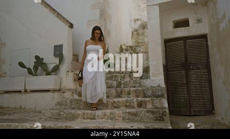 A young hispanic woman in a white dress walks down stone stairs in the charming old town of ostuni, puglia, italy, carrying a wicker bag. Stock Photo