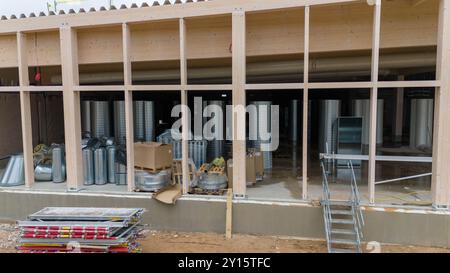 Construction site featuring a wooden structure with large windows. Inside, various metal ducts and equipment are visible, along with boxes and constru Stock Photo
