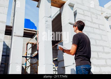 Man architect engineer checking house under construction comparing with blueprint plan on paper. Worker looks at window slot outside Stock Photo