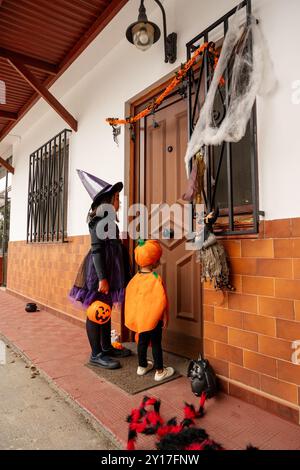 A girl in a witch costume is standing outside a house with a pumpkin in her hand. A little boy is also dressed in a costume and holding a pumpkin Stock Photo