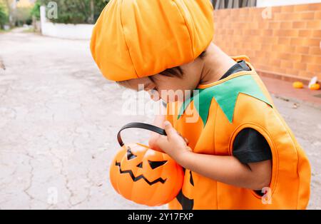 A little boy in a pumpkin costume holding a pumpkin bucket. The scene is set on a street, with a few other people in the background. Scene is playful Stock Photo