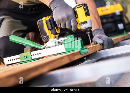 A carpenter is using a deckboard wrench and power drill to secure wooden boards on a construction site patio floor. Stock Photo