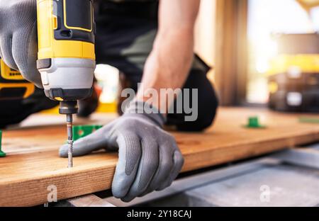 A construction worker in gloves focuses on driving a screw into wooden planks while using a power drill. The environment is bright and showcases ongoi Stock Photo