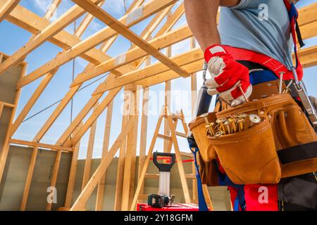 A construction worker holds tools in a leather belt while standing in a wooden frame building under construction, showcasing craftsmanship in progress Stock Photo
