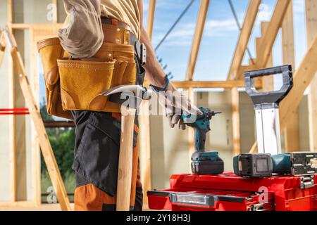 A construction worker stands at a building site, organizing tools and using a drill while wearing a tool belt. The sun shines brightly, illuminating t Stock Photo