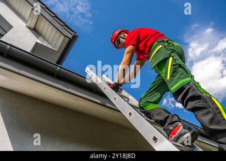 A maintenance worker in safety gear ascends a ladder to inspect and repair a roof gutter while the sun shines brightly overhead. Stock Photo