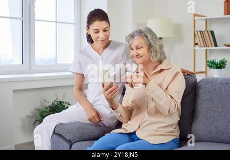 Young cheerful female nurse showing elderly woman how to use smartphone. Stock Photo