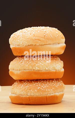 A stack of three hamburger buns with sesame seeds on a rustic wood surface and a light to dark warm background. Stock Photo