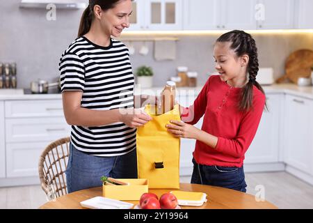 Mother with her daughter putting bottle of drink into lunch bag at wooden table in kitchen. Preparing for school Stock Photo
