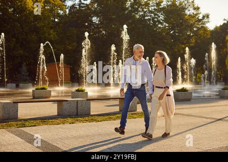 Senior happy couple enjoying urban calm walk in city near fountain, holding hands close, holiday Stock Photo