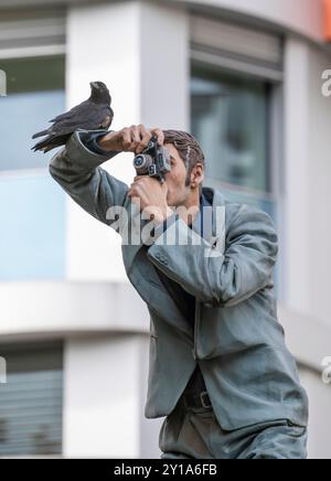 Real crow on a pillar saint, The Photographer, in front of the main station in Düsseldorf, one of 10 realistic, life-size sculptures of people on adve Stock Photo