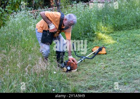 A mature man in protective clothing, and gloves with a trimmer. Mowing tall grass and the weeds with a trimmer. Selectively focus Stock Photo