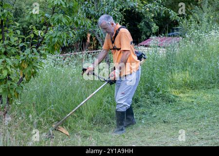 A mature man in protective clothing, and gloves with a trimmer. Mowing tall grass and the weeds with a trimmer. Selectively focus Stock Photo