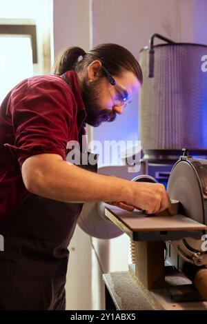 Carpenter in woodworking shop wearing protection goggles while using disc sander to avoid injury. Cabinetmaker in joinery using professional grinding machine with safety gear on to prevent accidents Stock Photo