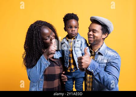 Cheerful african american mom and dad showing thumbs up sign and holding their infant son, grateful family bonding against yellow background. Positive husband and wife give like symbol. Stock Photo