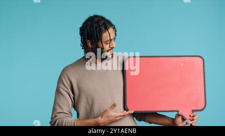 Joyful BIPOC man holding red speech bubble sign of empty space for text. Upbeat Middle Eastern person presenting thought bubble cardboard used as advertisement, isolated over blue studio background Stock Photo