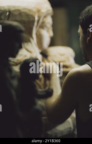 The silhouette of a craftsman seen carving wood with detailed focus on statues in a dim workshop Stock Photo