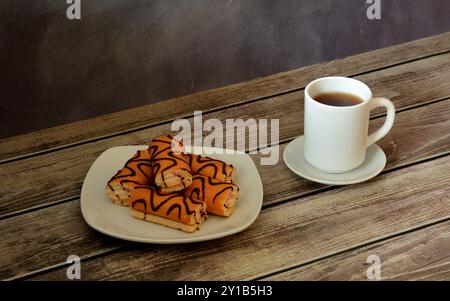 Mini sponge rolls with cream and chocolate icing on a white plate, next to a cup of hot drink on a wooden table. Close-up. Stock Photo