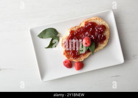 Slice of bread with raspberry jam decorated with fruit berries and leaves on white ceramic plate on white wooden table. Top view. Stock Photo