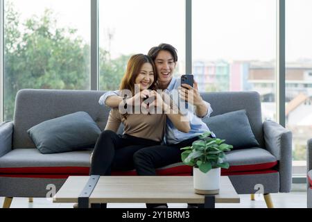 Young asian couple sitting on a couch, making a heart shape with their hands via remote communication through smartphone, in a bright living room. Stock Photo