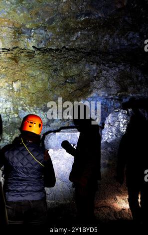 A group of people are standing in a dark cave, with one person wearing an orange helmet. The cave is illuminated by a light, and the group appears to Stock Photo